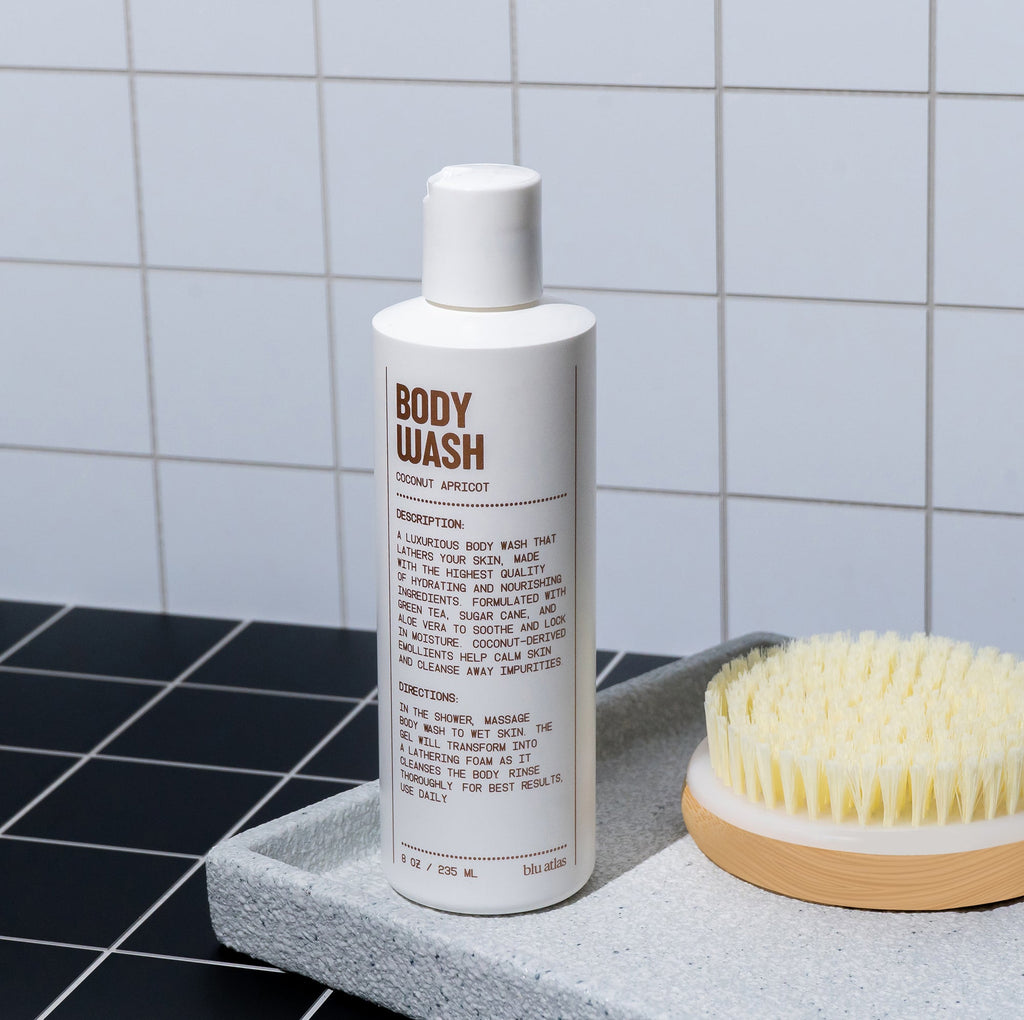 Body wash bottle with a brush on a gray surface against a tiled wall.