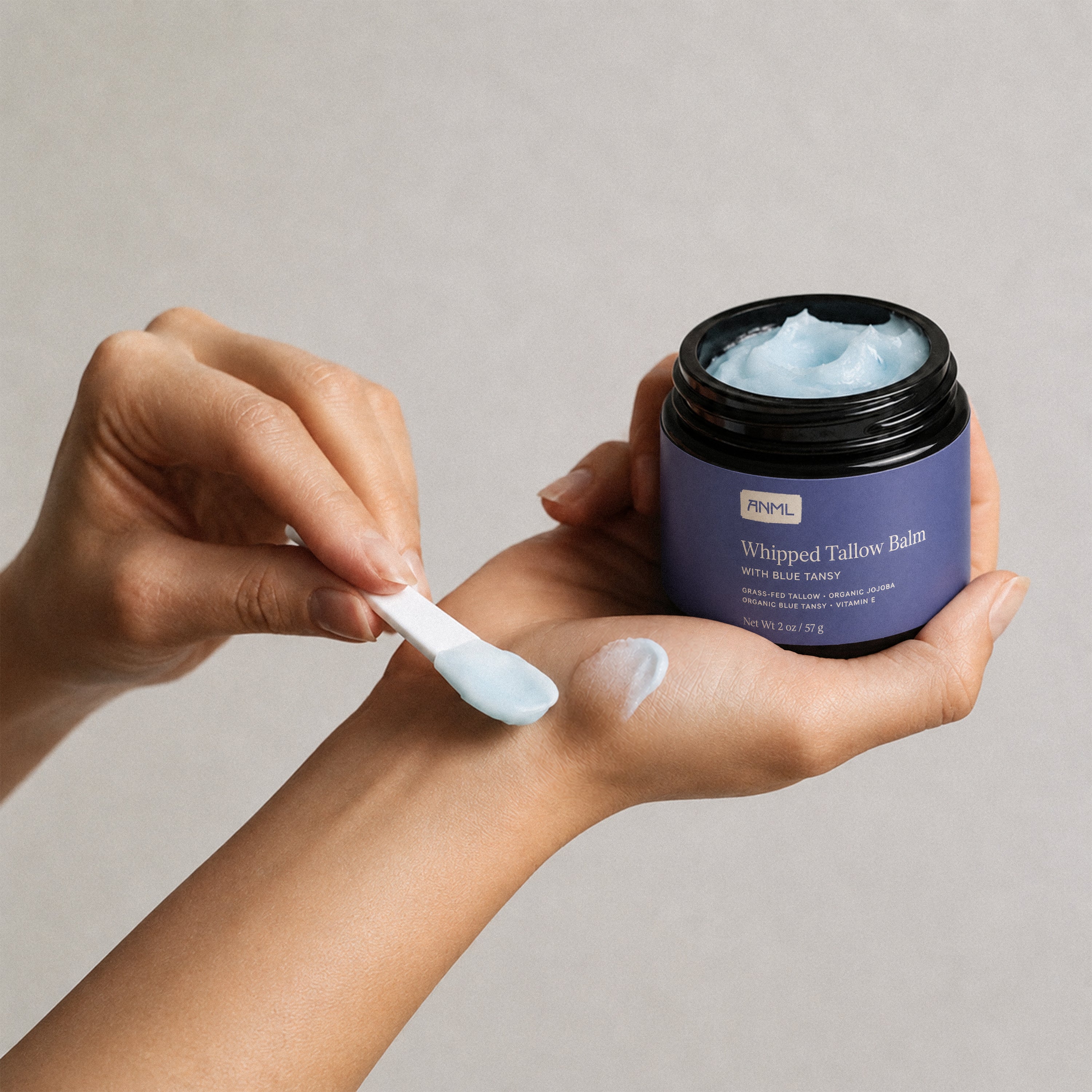 Person applying whipped tallow balm from a jar with a spoon on a neutral background
