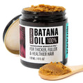 A jar of Unrefined Banana Oil hair treatment with its lid off, showing the thick brown paste inside. A small wooden spoon in the foreground holds more of the brown oil, set against a clean white background.