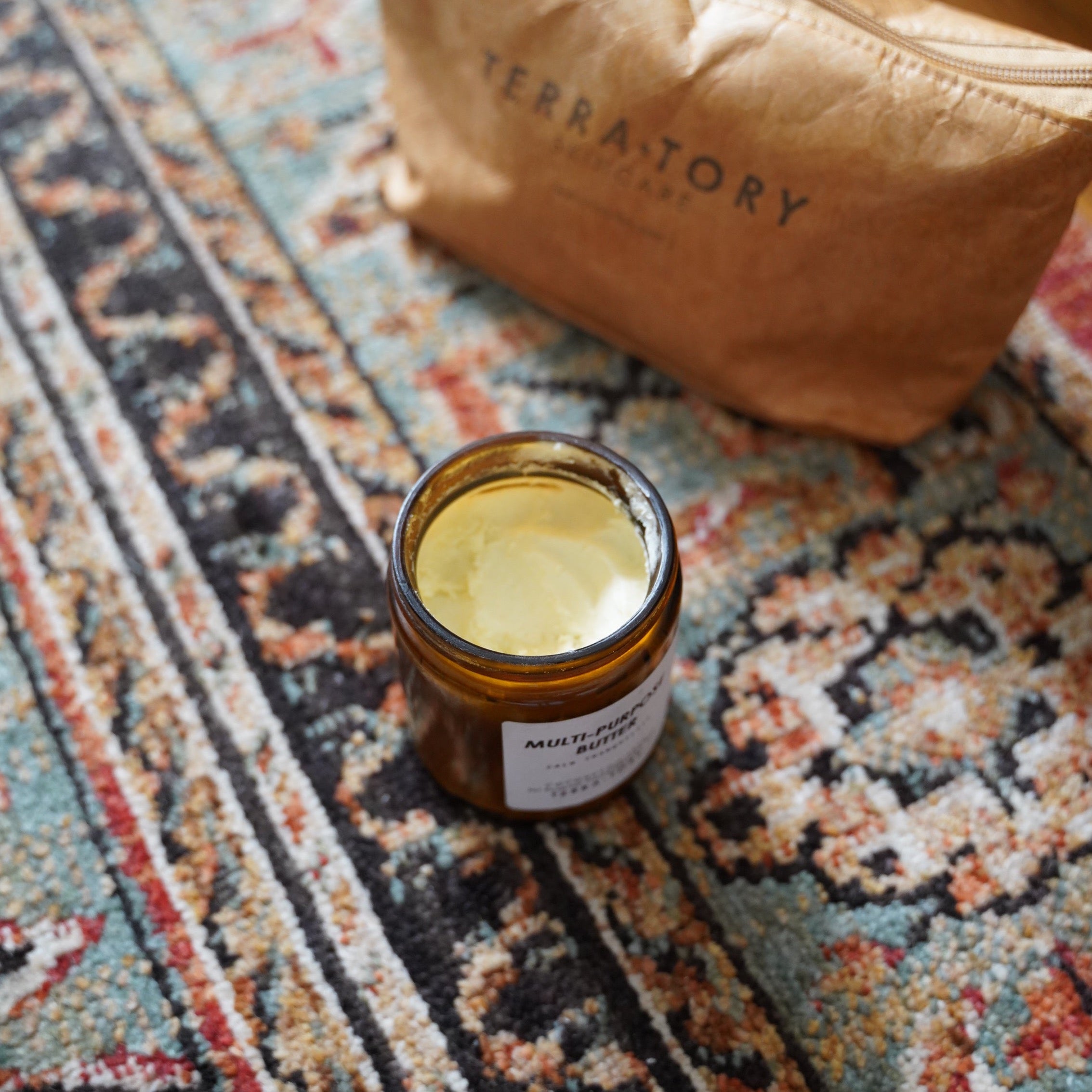 Jar of cream on a patterned rug with 'Terra Tory' bag in the background