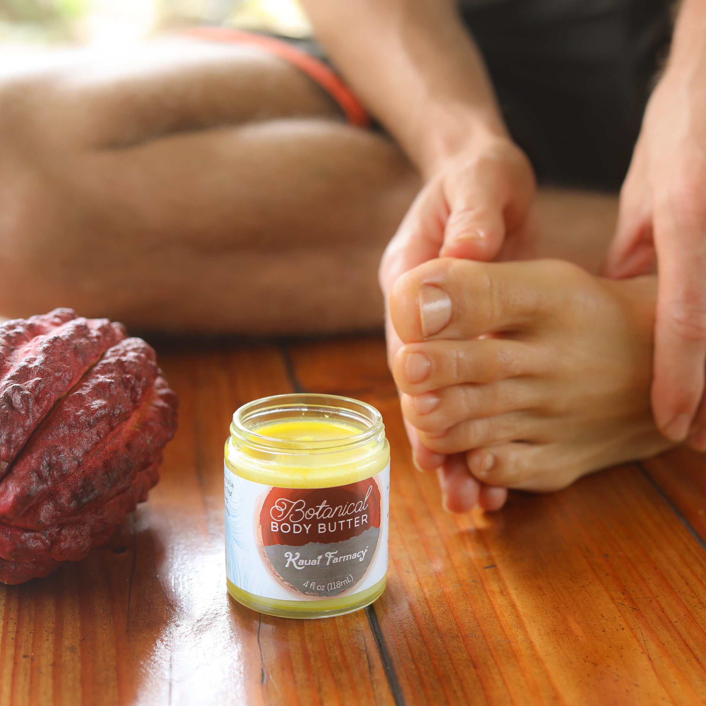 Jar of Botanical Body Butter on a wooden surface with a person applying it to their hand.